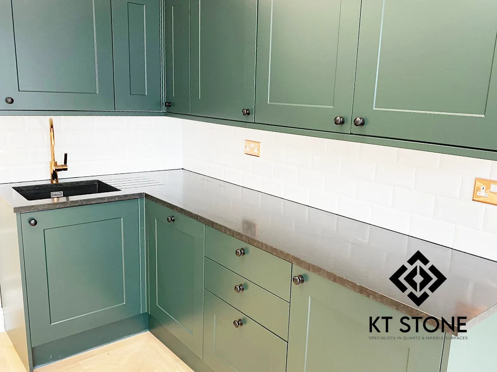 L-shaped dark quartz worktop with green shaker cabinets and a white tiled splashback, from a KT Stone project in London.